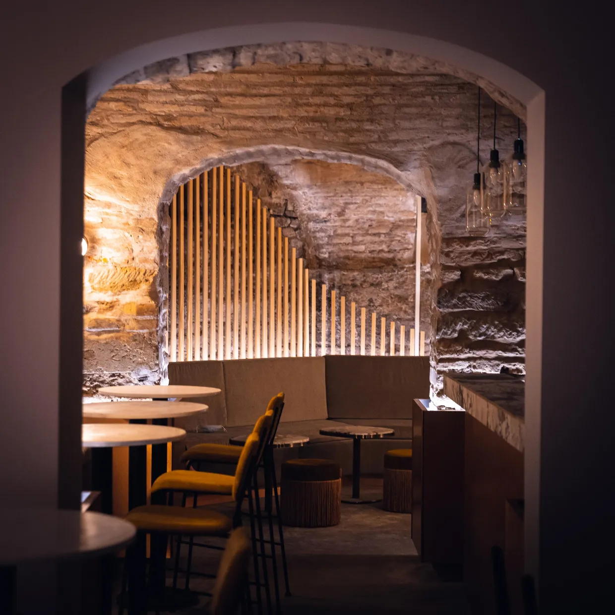Un espace intérieur avec des murs en pierre brute, des arches et des lumières, contenant des tables rondes et des chaises autour de banquettes