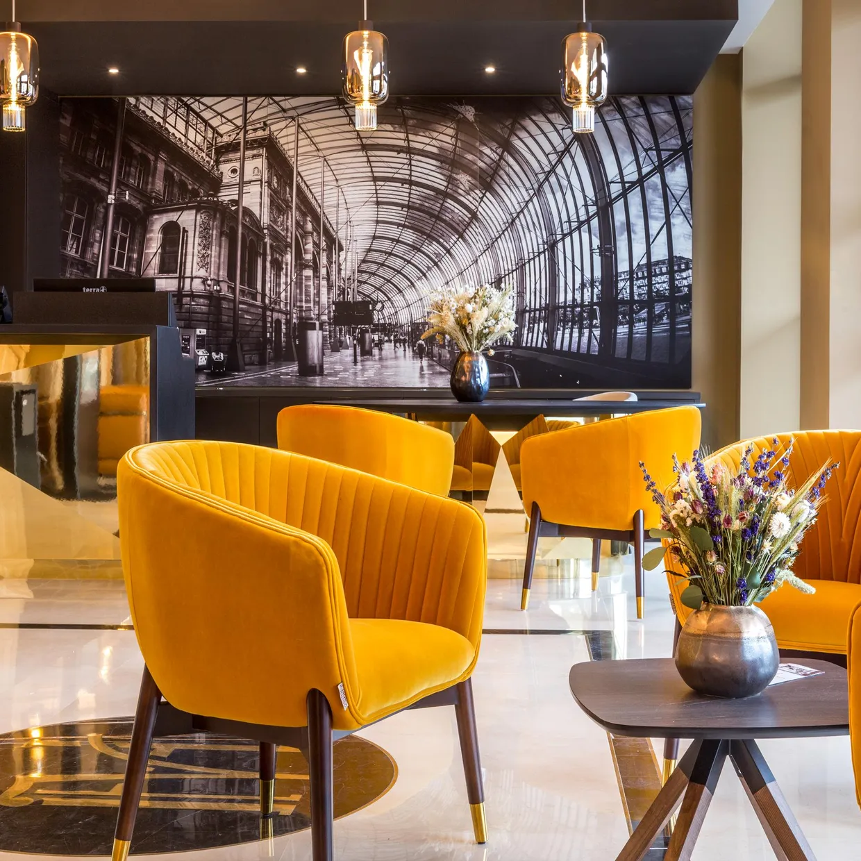 Un espace intérieur avec des chaises jaunes, une table basse avec un vase de fleurs, un comptoir d'accueil et une grande photographie en noir et blanc d'une gare