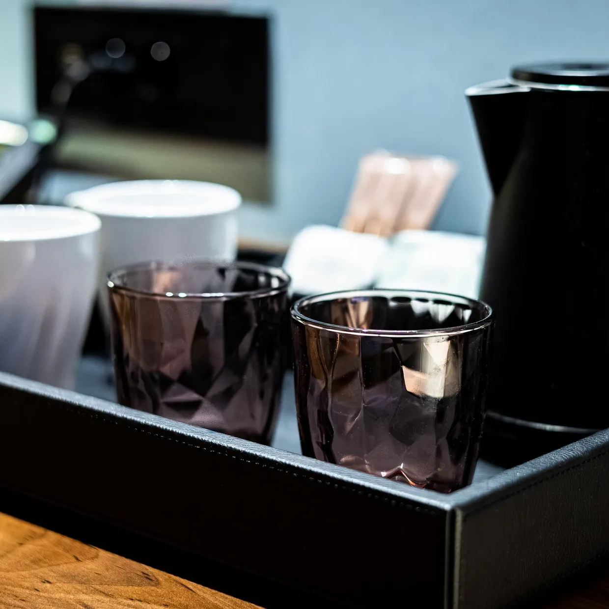 Un plateau avec une bouilloire noire, deux tasses blanches et deux verres en verre fumé sur une table en bois
