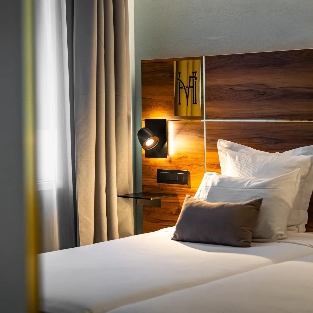 Lit avec une tête de lit en bois, des oreillers blancs et un coussin gris, éclairé par une lampe de chevet