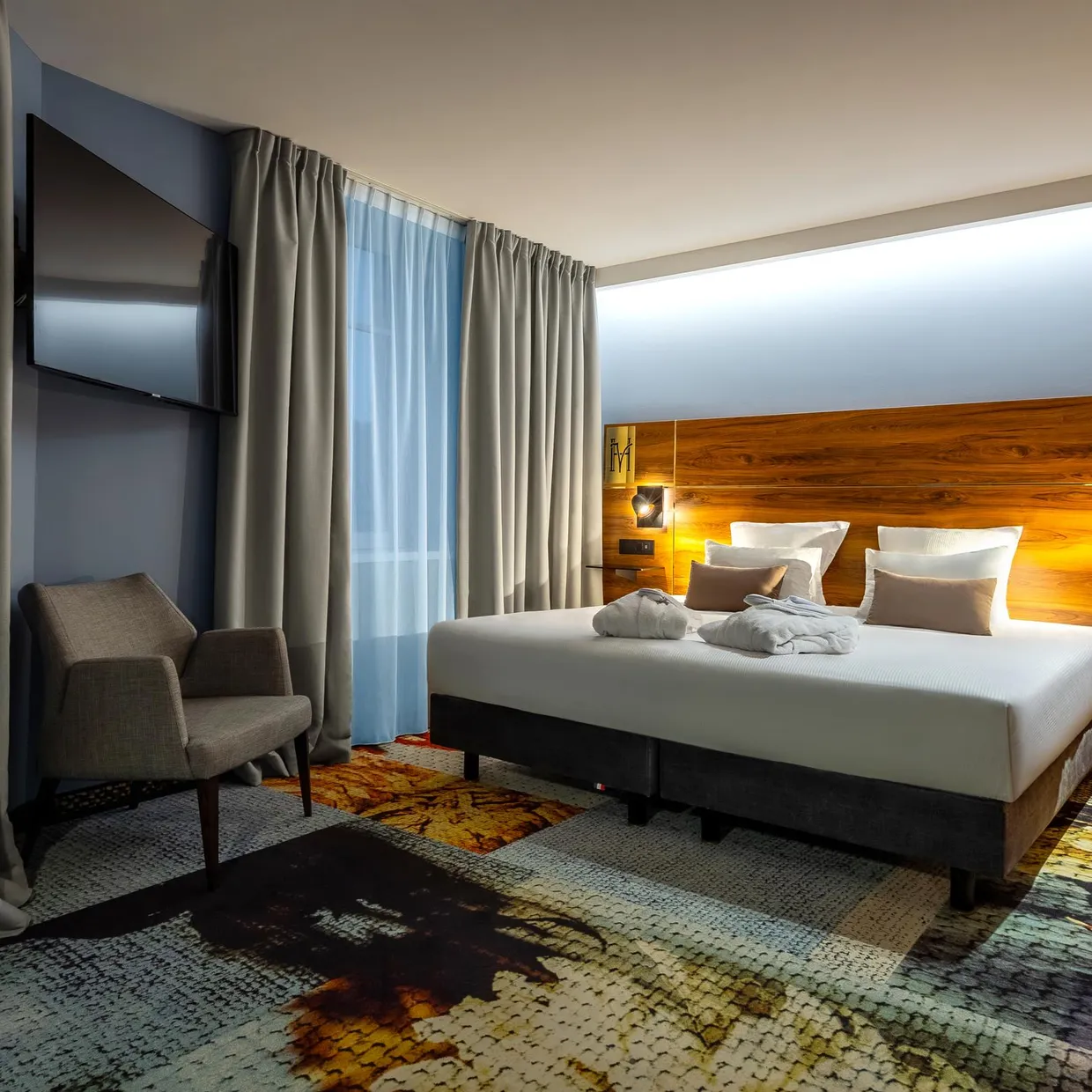 Chambre d'hôtel avec un lit double, deux fauteuils, une tête de lit en bois et des rideaux gris