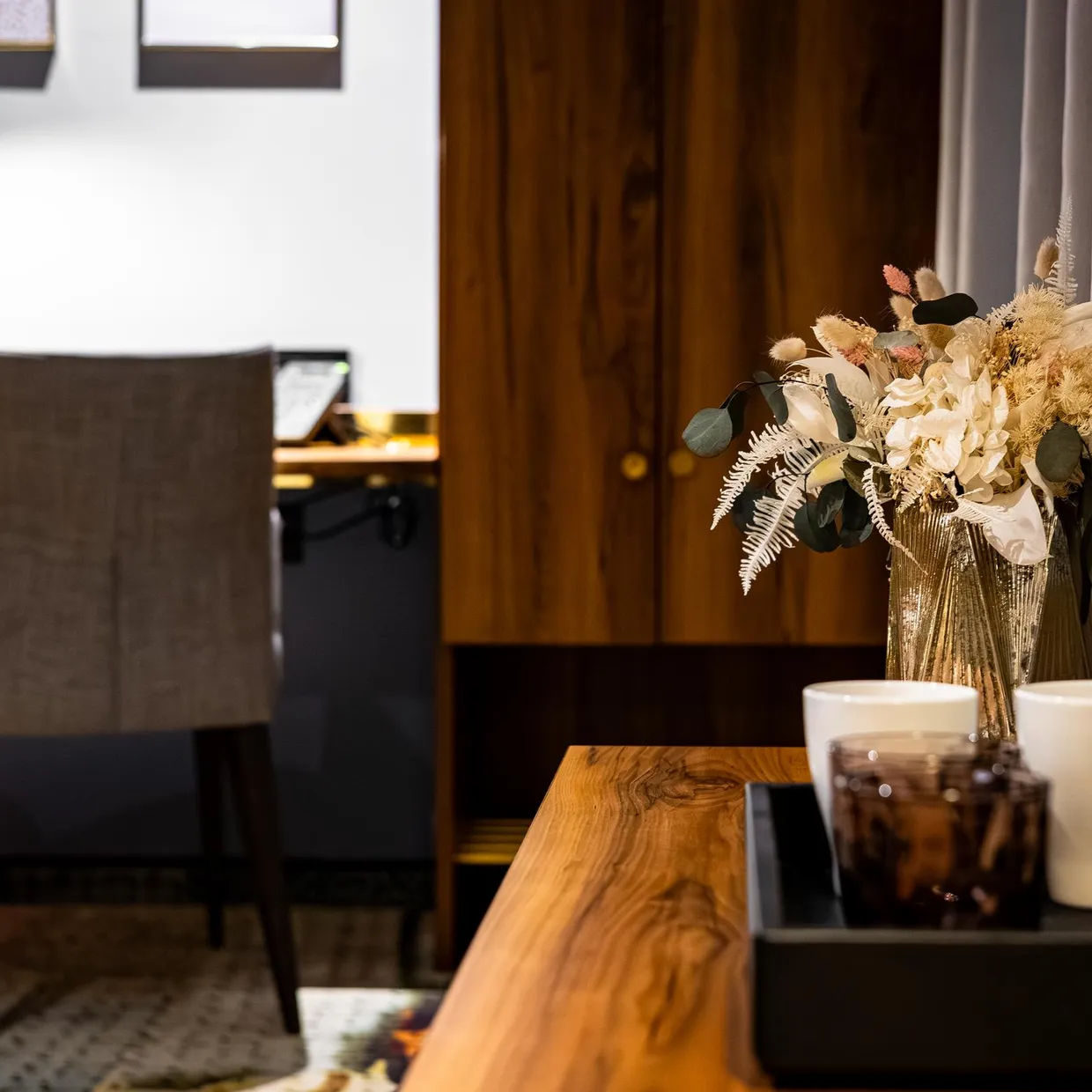 Un vase avec des fleurs blanches et des feuilles vertes et brunes sur une table en bois, avec une chaise en tissu et un bureau en arrière-plan