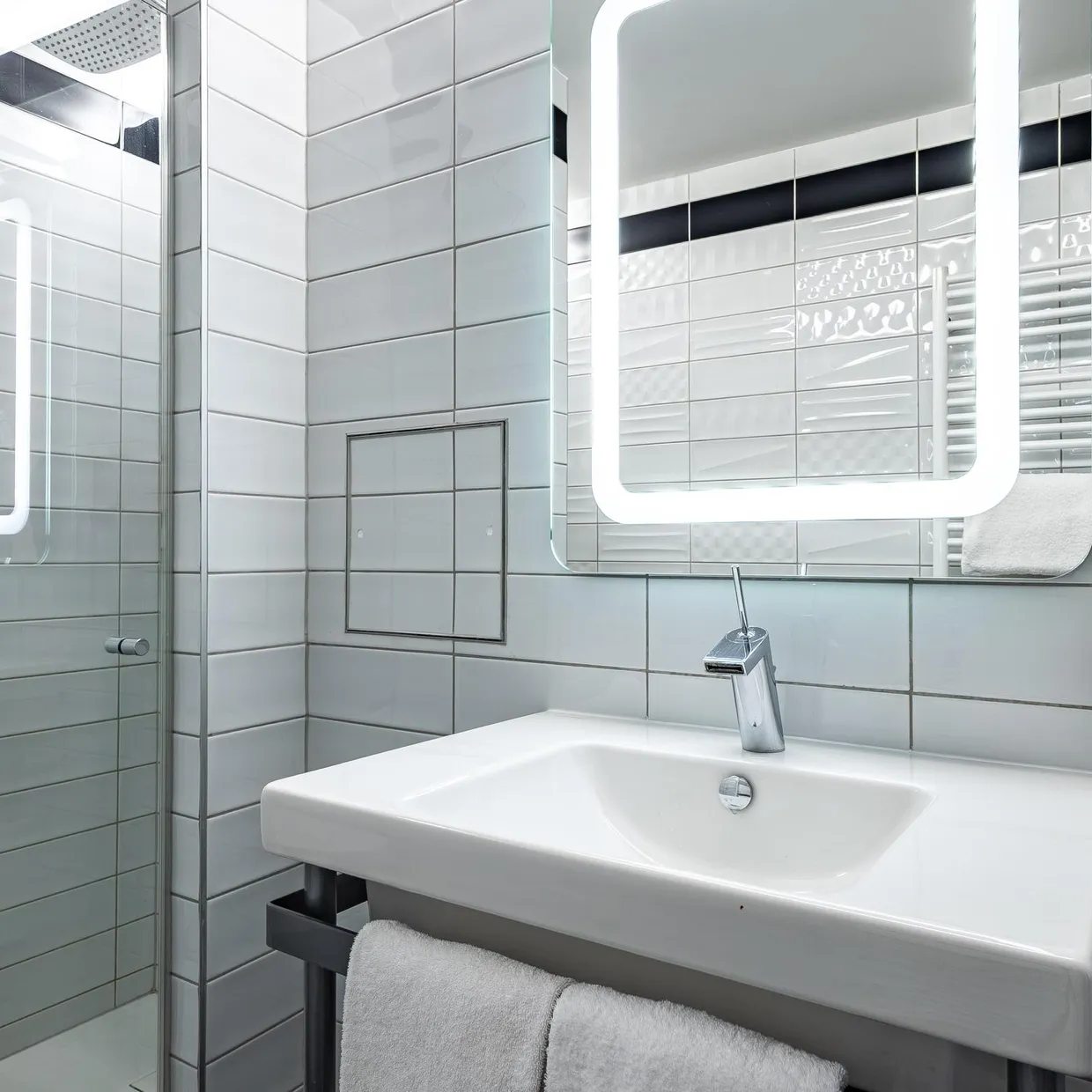 Un lavabo blanc avec un robinet, des serviettes suspendues, un distributeur de savon et un miroir éclairé dans une salle de bain carrelée de blanc
