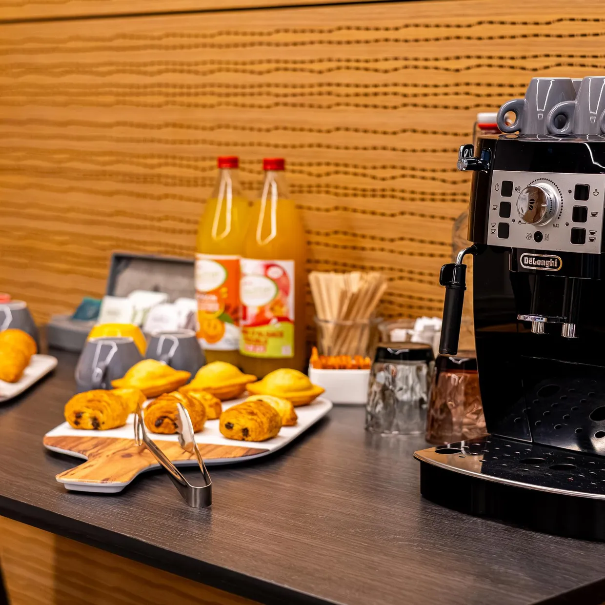 Table avec une machine à café, des tasses, des bouteilles de jus d'orange, des oranges et des pâtisseries