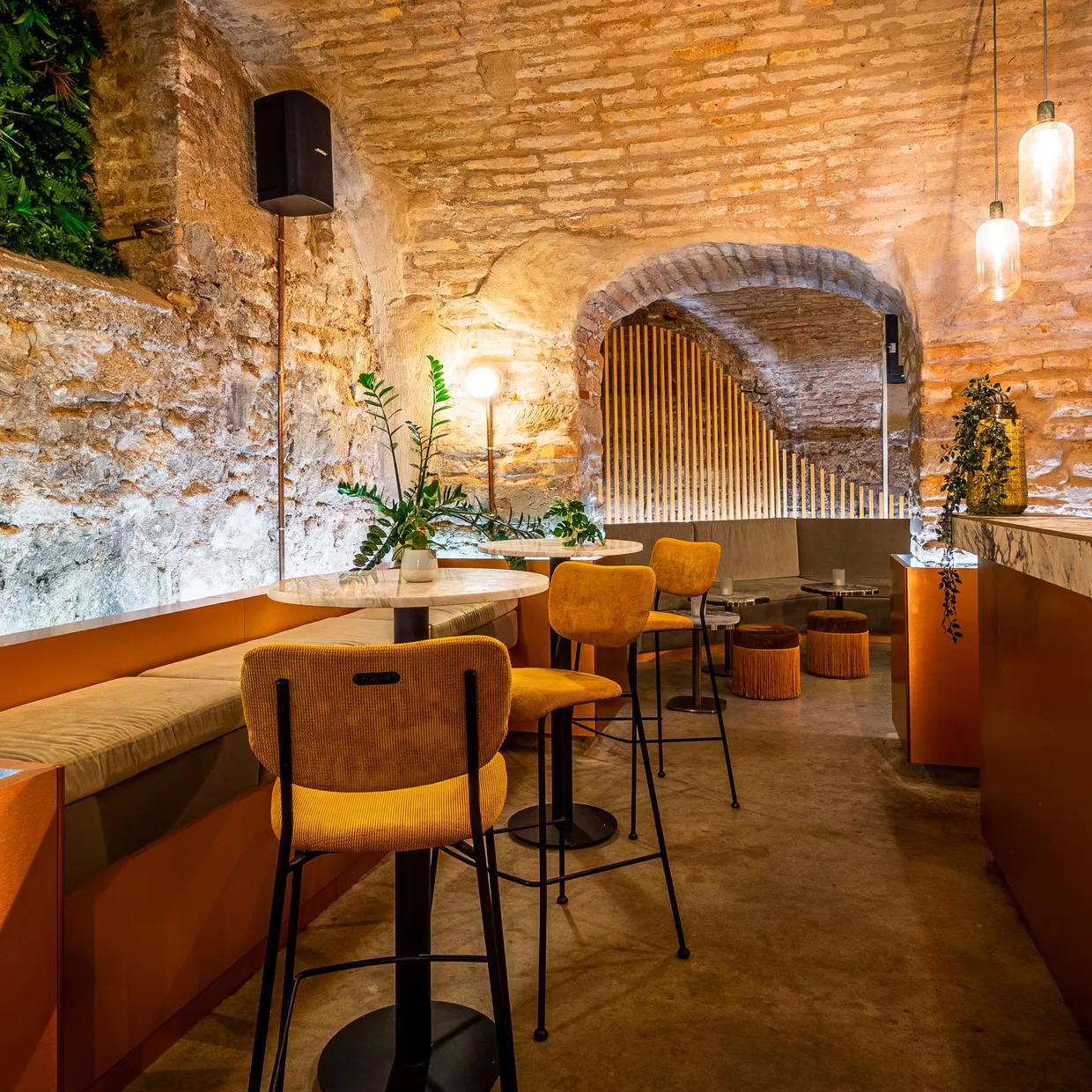 Un espace intérieur avec des murs en briques apparentes, des sièges orange, des tables et des plantes, éclairé par des lampes suspendues