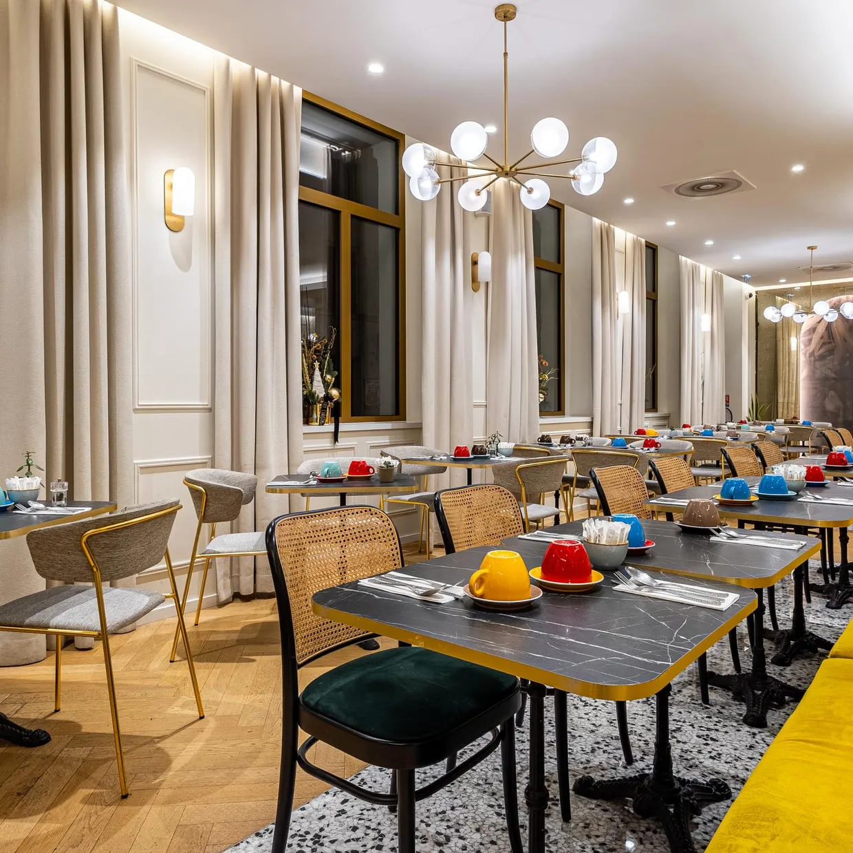 Un café élégant avec des tables rondes, des chaises et un canapé jaune, éclairé par des suspensions modernes et des appliques murales