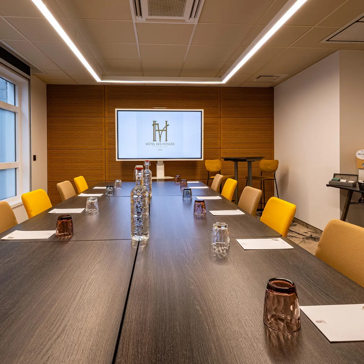 Salle de réunion avec une table en bois, des chaises jaunes, des verres et des carafes d'eau, un écran avec un logo et une table avec des rafraîchissements
