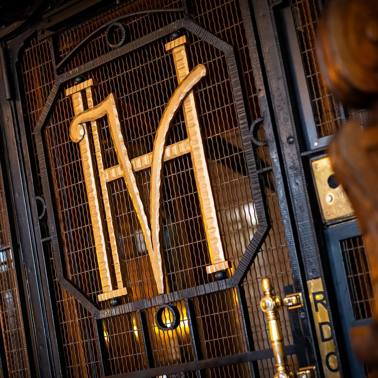 Grille métallique avec les lettres "M" et "A" en relief, éclairée par des lumières dorées, dans un cadre architectural sombre
