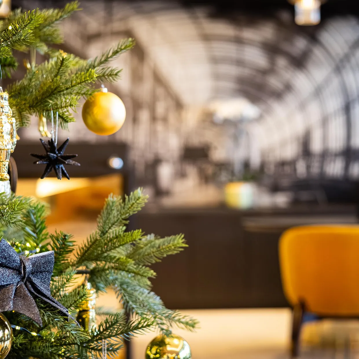 Un sapin de Noël décoré avec des ornements dorés et noirs, placé dans un intérieur avec des chaises jaunes et un mur en briques