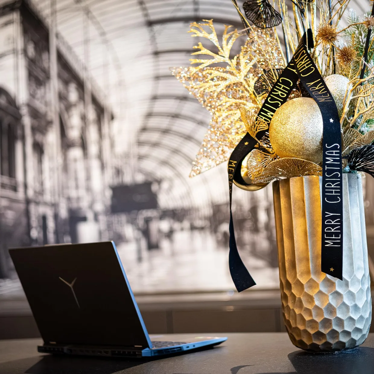 Un vase décoré avec des éléments festifs et un ruban "Merry Christmas" est posé sur une table à côté d'un ordinateur portable devant une grande verrière
