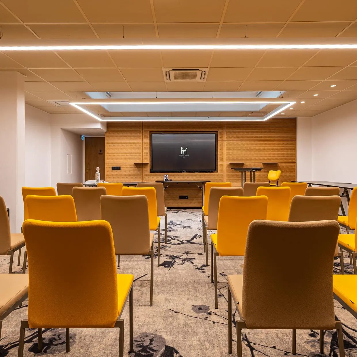 Salle de réunion avec des chaises orange et des tables, un écran et un coin café en fond
