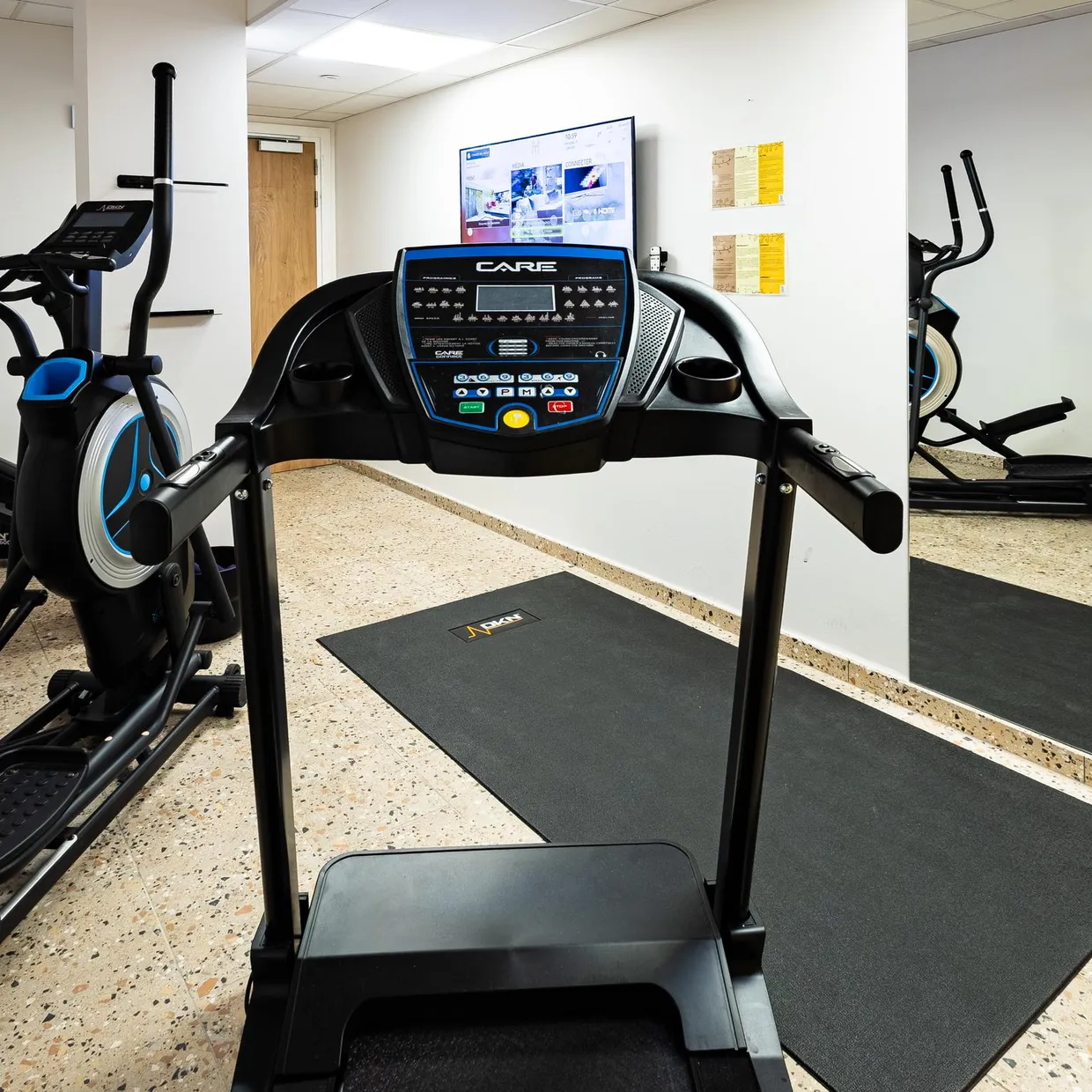 Salle de sport avec plusieurs appareils de cardio-training, dont un tapis de course et deux vélos elliptiques, placés sur des tapis de sol