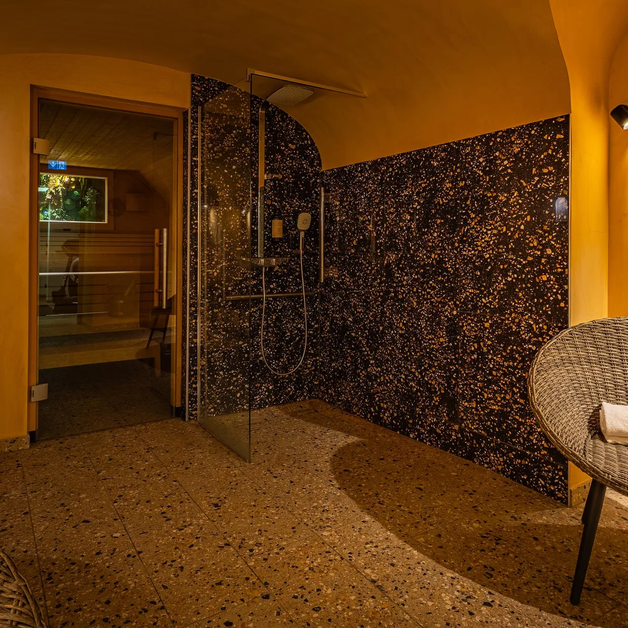 Salle de douche avec paroi en mosaïque noire et dorée, deux chaises en rotin et une porte ouverte sur une pièce adjacente