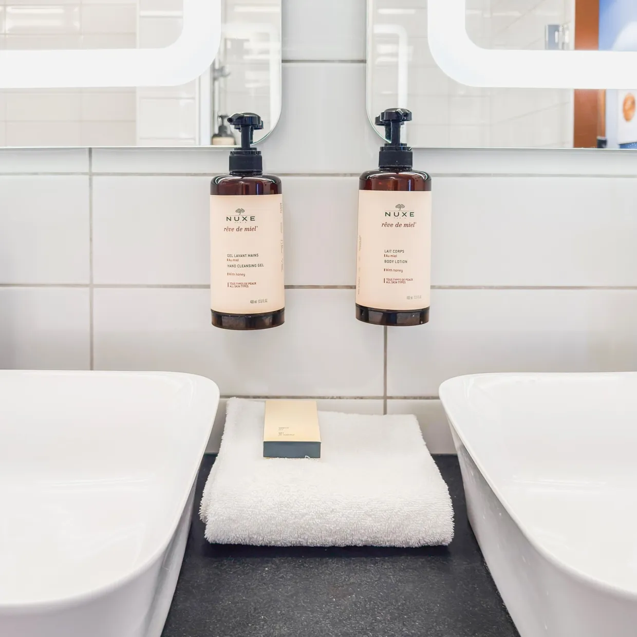Deux distributeurs de produits Nuxe "rêve de miel" suspendus entre deux lavabos avec serviettes et savon