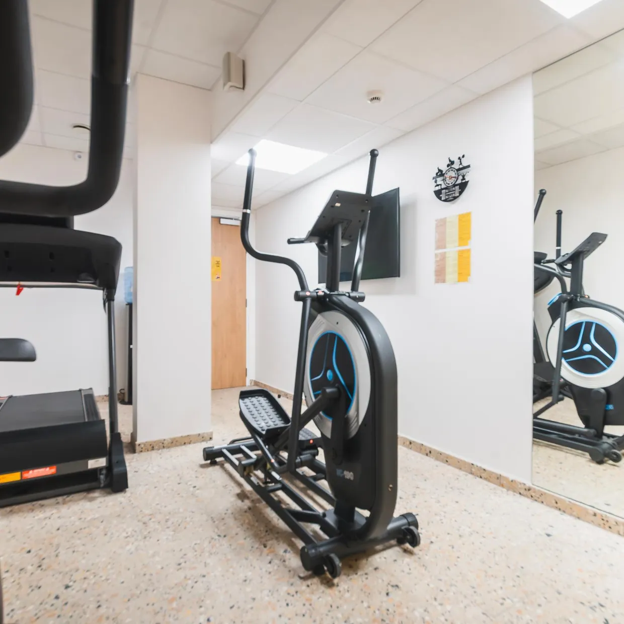 Salle de sport avec plusieurs appareils de cardio-training, dont un tapis de course et des vélos elliptiques, devant un miroir