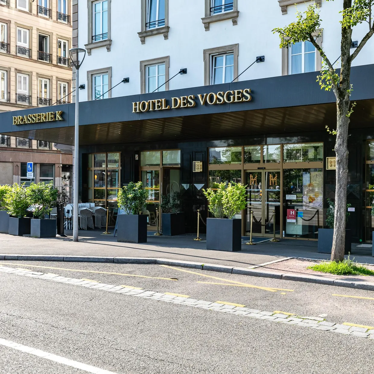 Entrée d'un hôtel nommé "Hotel des Vosges" avec une brasserie adjacente, située dans une rue urbaine avec des plantes en pots devant l'entrée