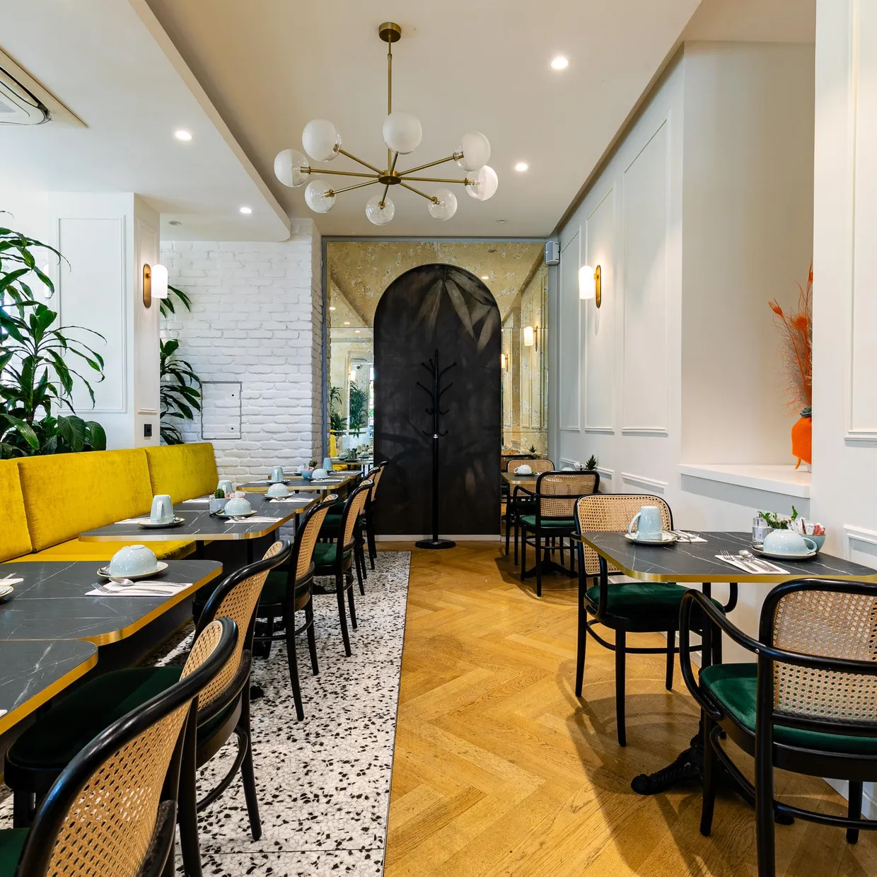 Restaurant intérieur avec tables noires, chaises en rotin et banquette jaune, décoré de plantes vertes et éclairé par un lustre à globes
