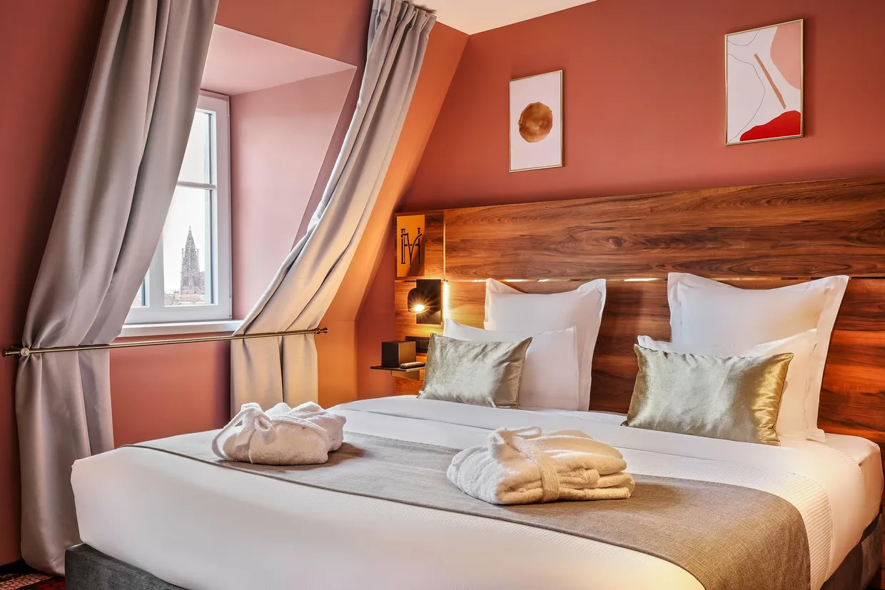 Chambre avec un lit double, des oreillers et des couvertures, une tête de lit en bois, des rideaux gris et une fenêtre avec une vue sur une tour