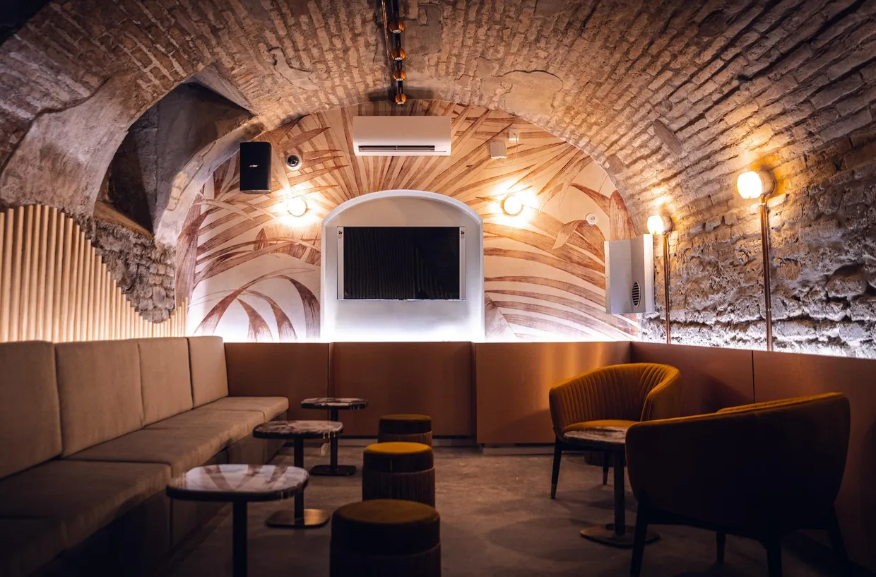 Salle souterraine avec arches en pierre, canapé beige, fauteuils jaunes et écran au fond