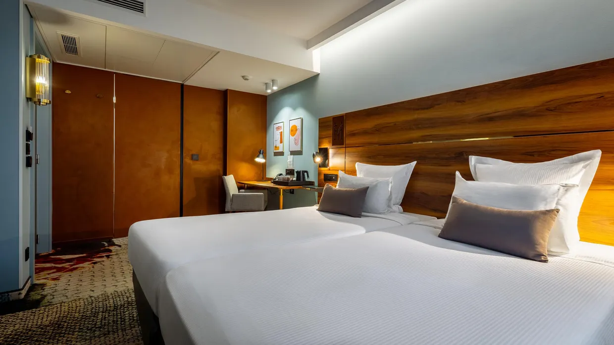 Chambre d'hôtel avec deux lits simples, une tête de lit en bois, un bureau avec une chaise et des œuvres d'art au mur