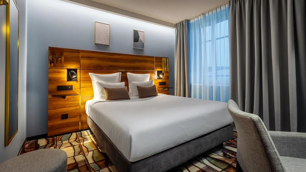 Chambre d'hôtel avec un grand lit double, des oreillers blancs et des coussins bruns, une tête de lit en bois avec éclairage intégré, des rideaux bleus et une fenêtre laissant entrer la lumière du jour