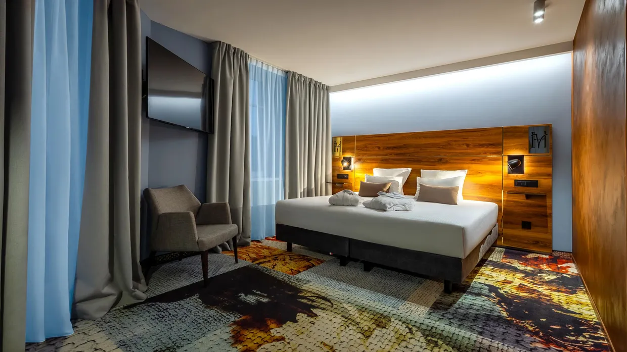 Chambre d'hôtel avec un lit double, deux fauteuils, une tête de lit en bois et des rideaux gris