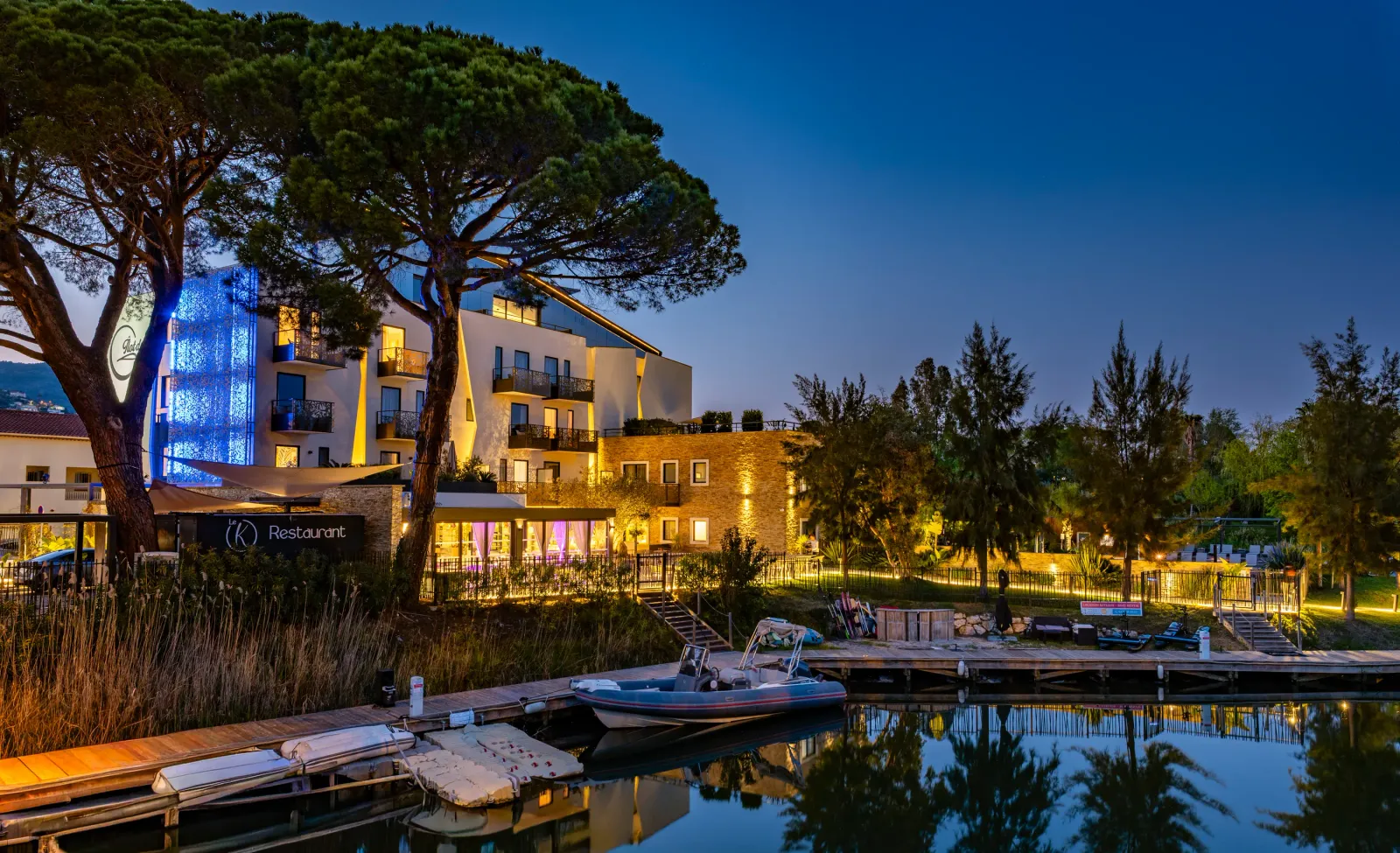Bâtiment moderne éclairé avec un restaurant au rez-de-chaussée, entouré d'arbres et situé près d'un plan d'eau avec des bateaux amarrés