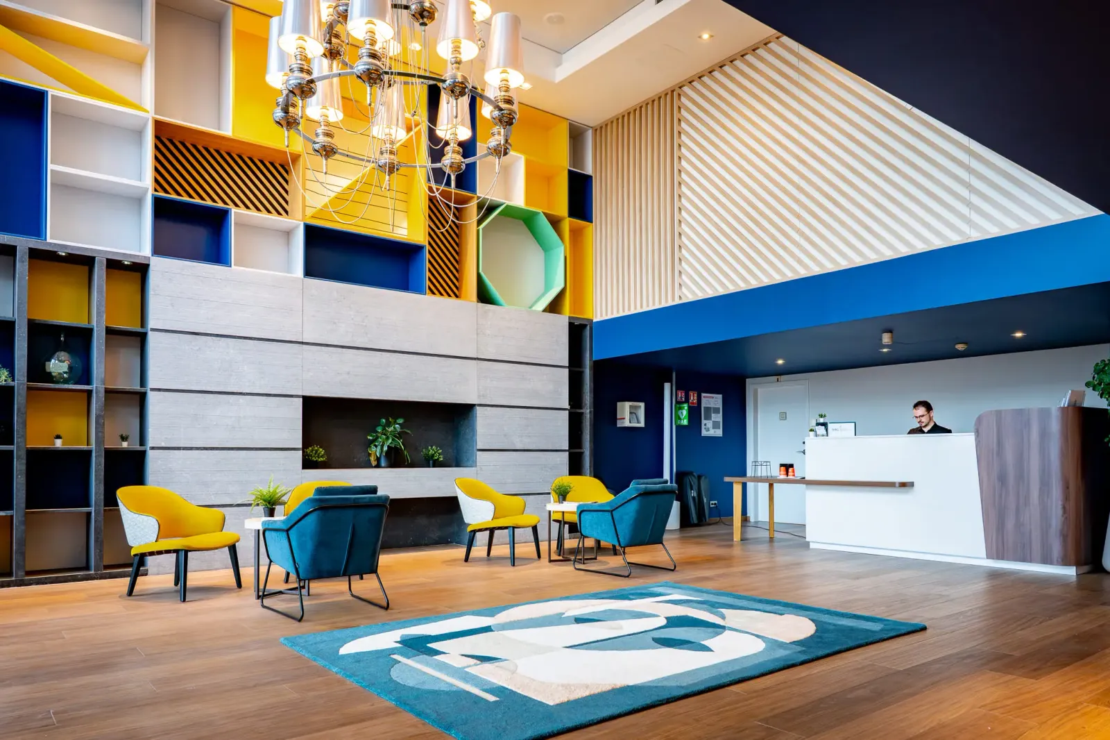 Réception d'hôtel moderne avec des chaises colorées, une table basse, un comptoir d'accueil et un lustre suspendu