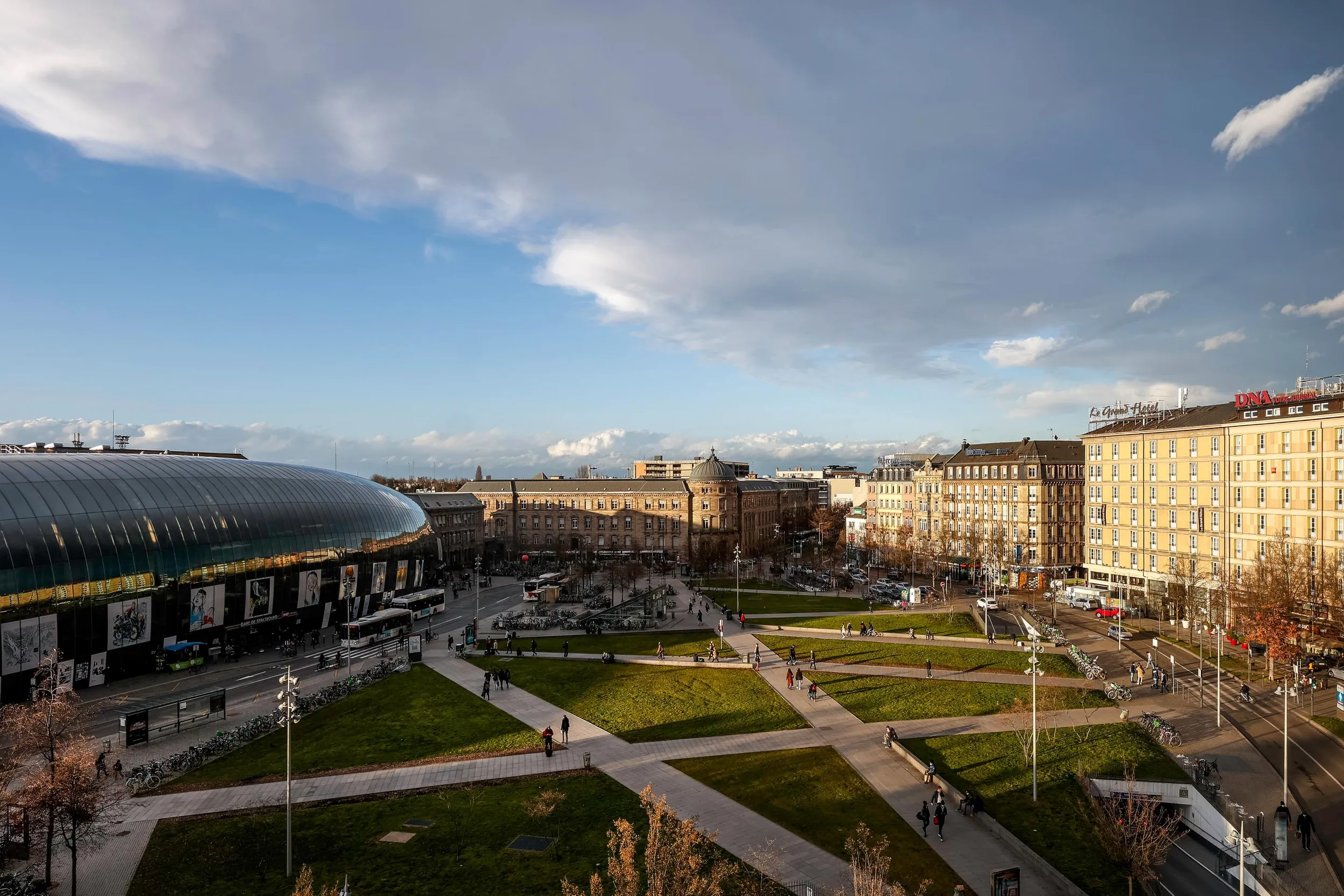 Vue panoramique d'une place urbaine avec un parc central, des bâtiments historiques et modernes, et une structure architecturale distinctive en forme de dôme