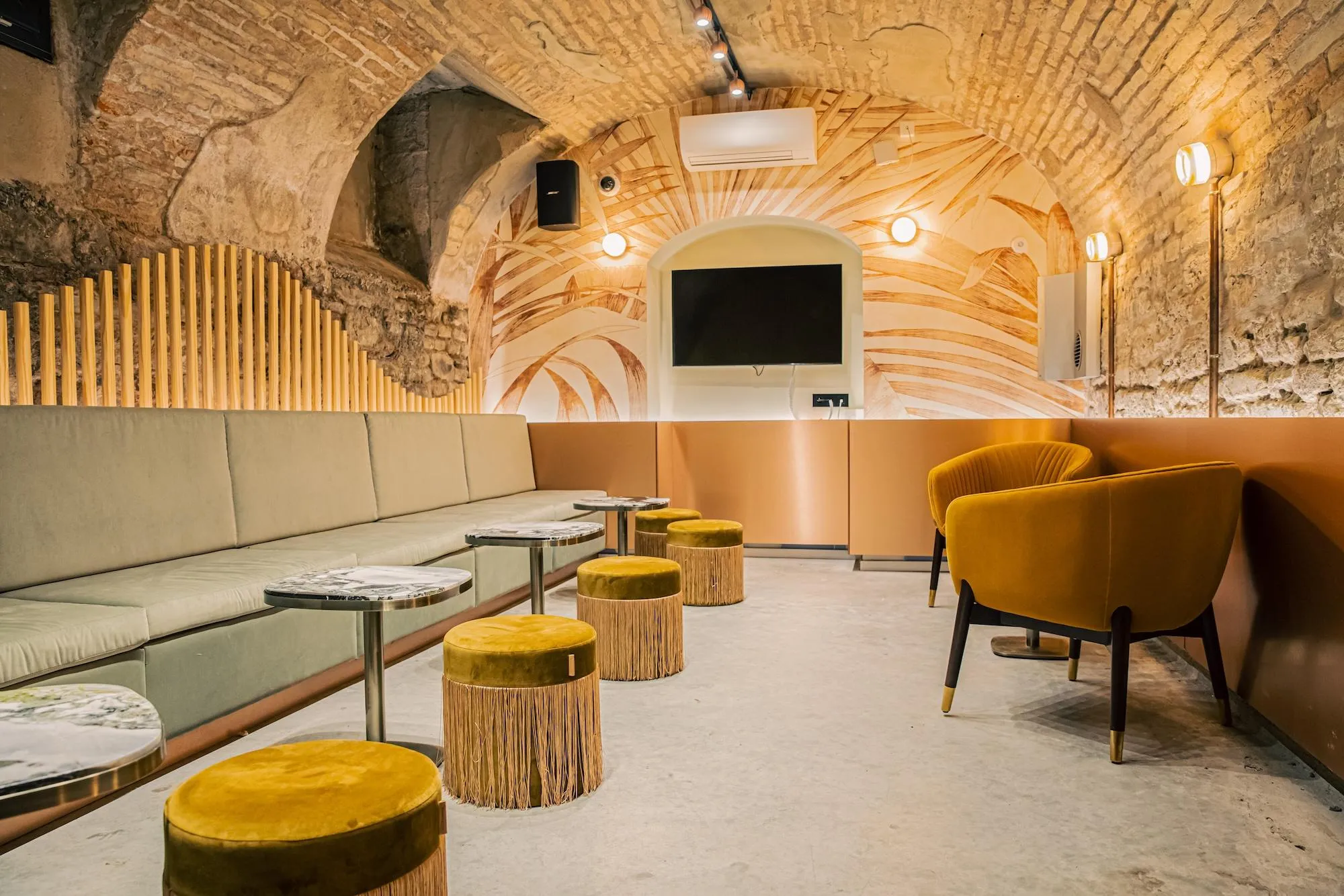 Un espace intérieur avec des arches en pierre, un plafond décoré de motifs en bois, un canapé beige, des tabourets et des fauteuils en velours jaune, ainsi qu'une télévision montée au mur