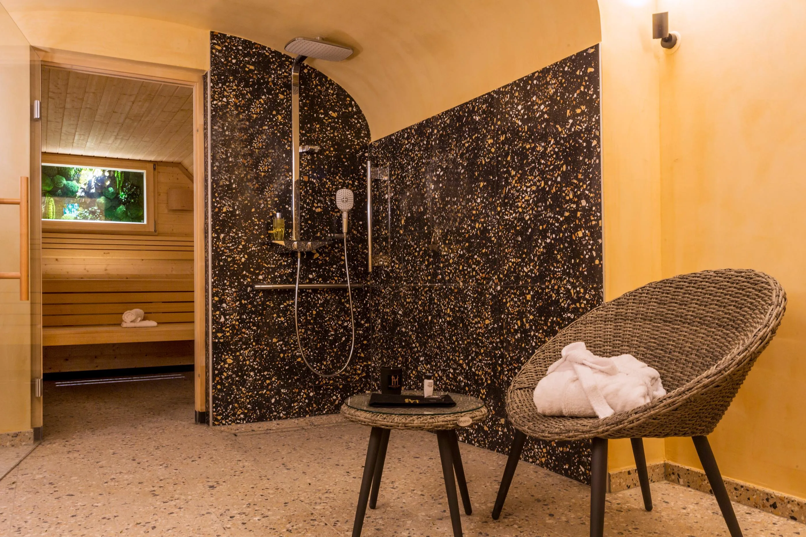 Salle de bain avec douche murale à paroi noire et dorée, chaise en osier avec serviette blanche et table basse