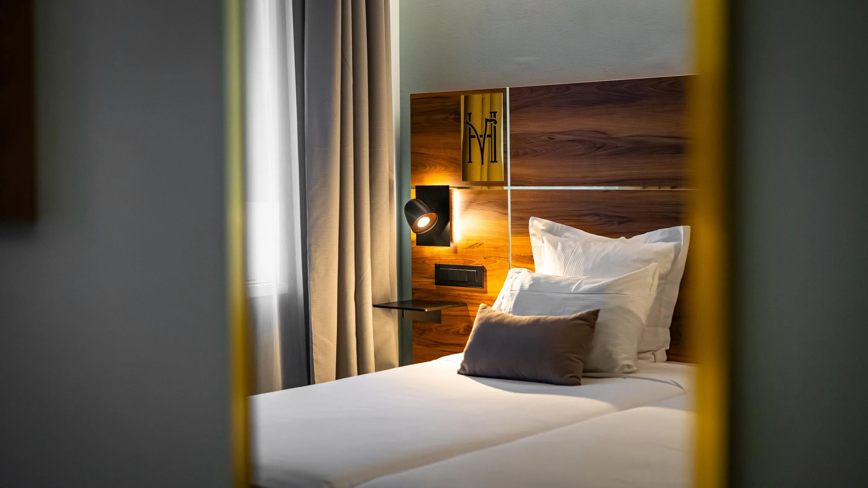 Lit avec une tête de lit en bois, des oreillers blancs et un coussin gris, éclairé par une lampe de chevet