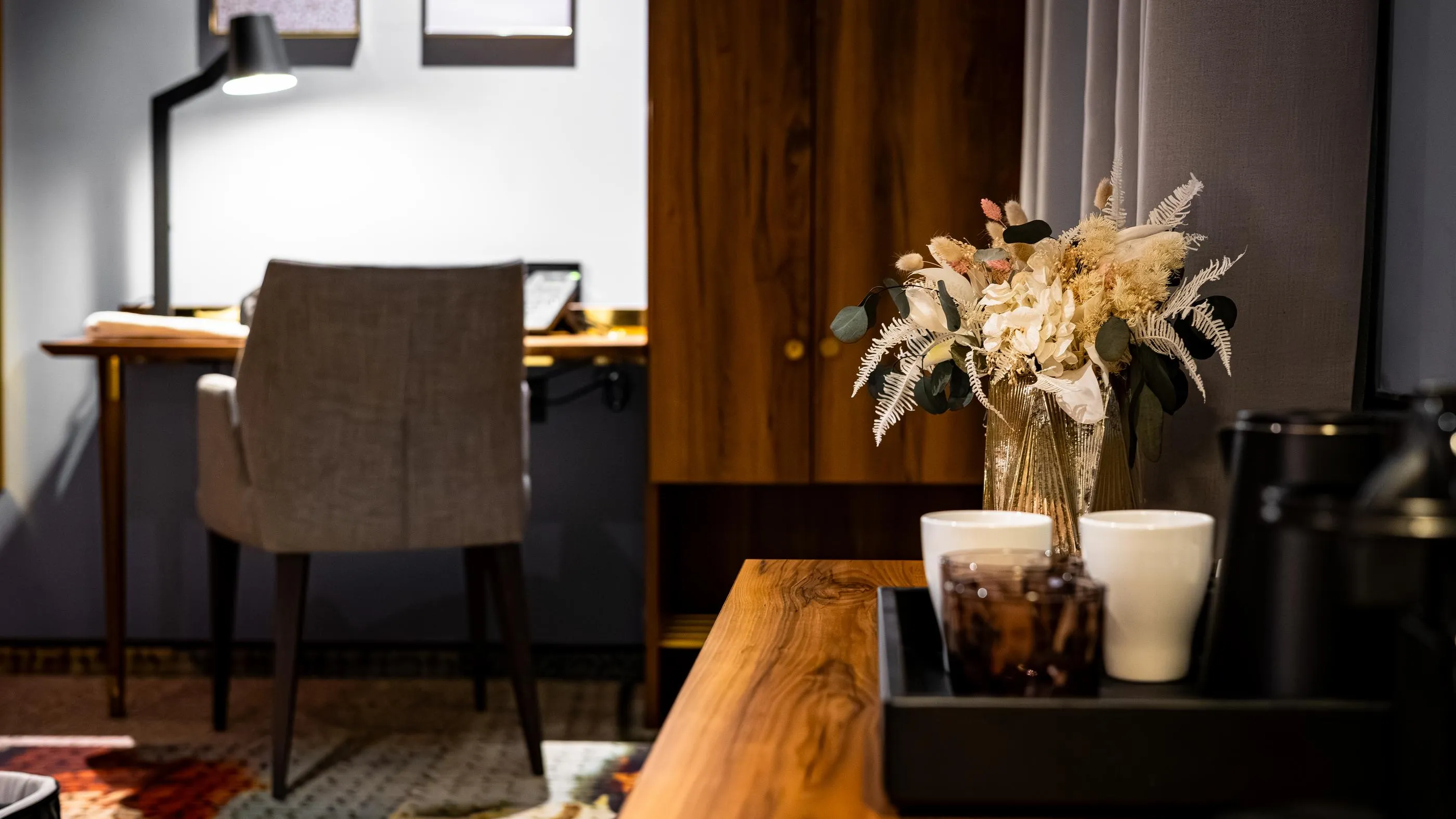 Un vase avec des fleurs blanches et des feuilles vertes et brunes sur une table en bois, avec une chaise en tissu et un bureau en arrière-plan