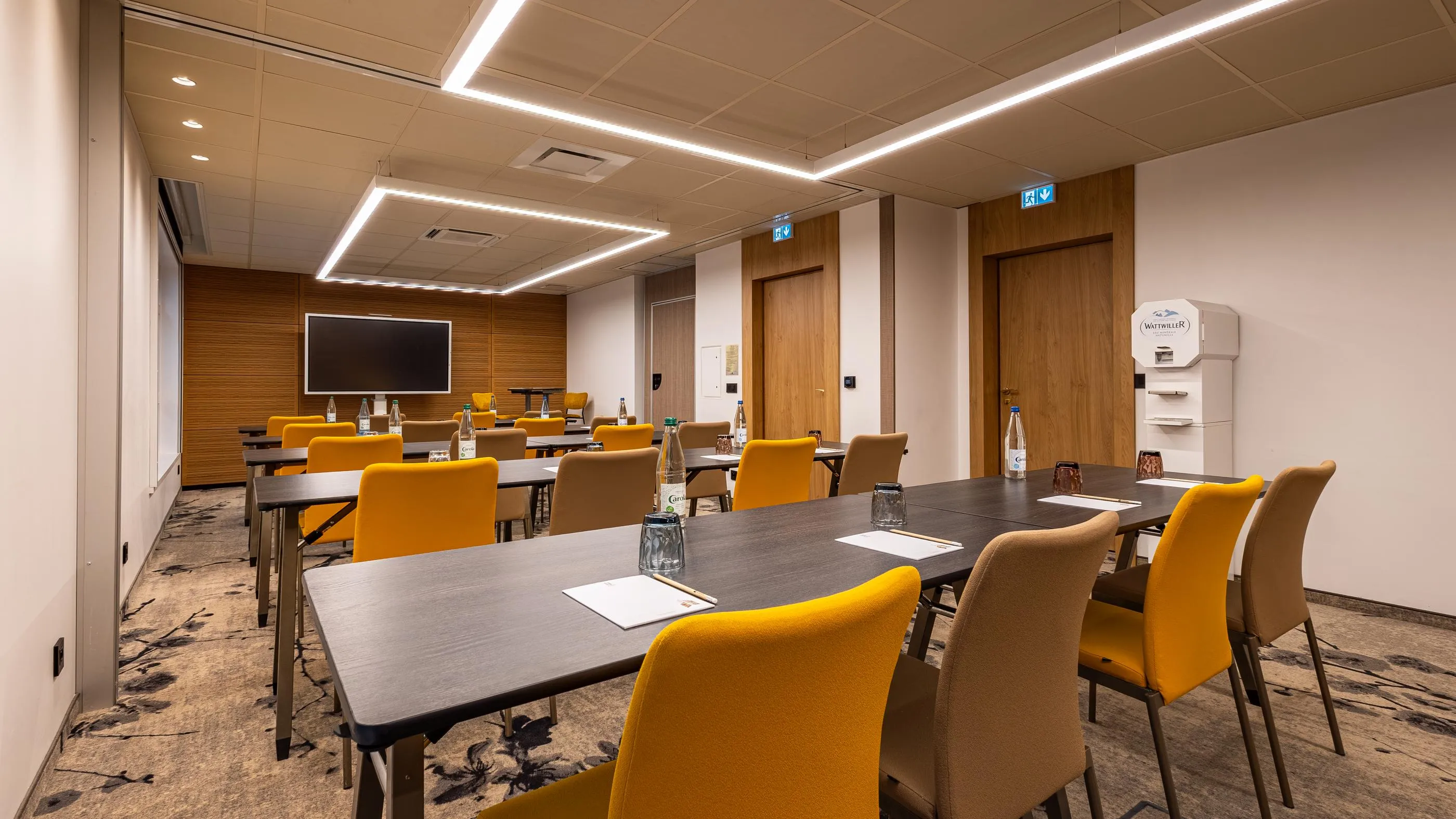 Salle de réunion avec tables et chaises, écran au fond, distributeur d'eau et bouteilles sur les tables