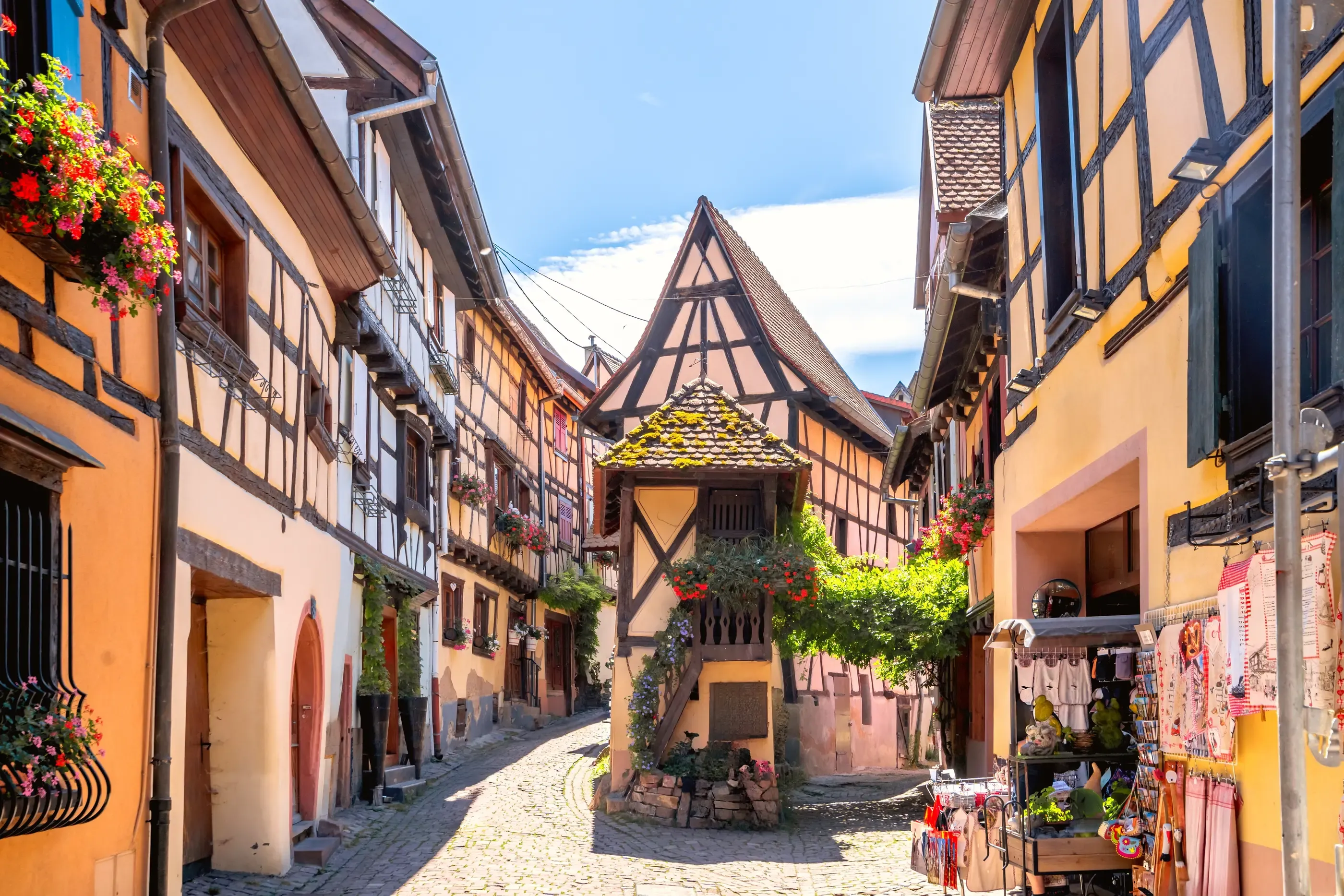 Rue pavée bordée de maisons à colombages colorées avec des fleurs suspendues et un petit kiosque en bois au centre