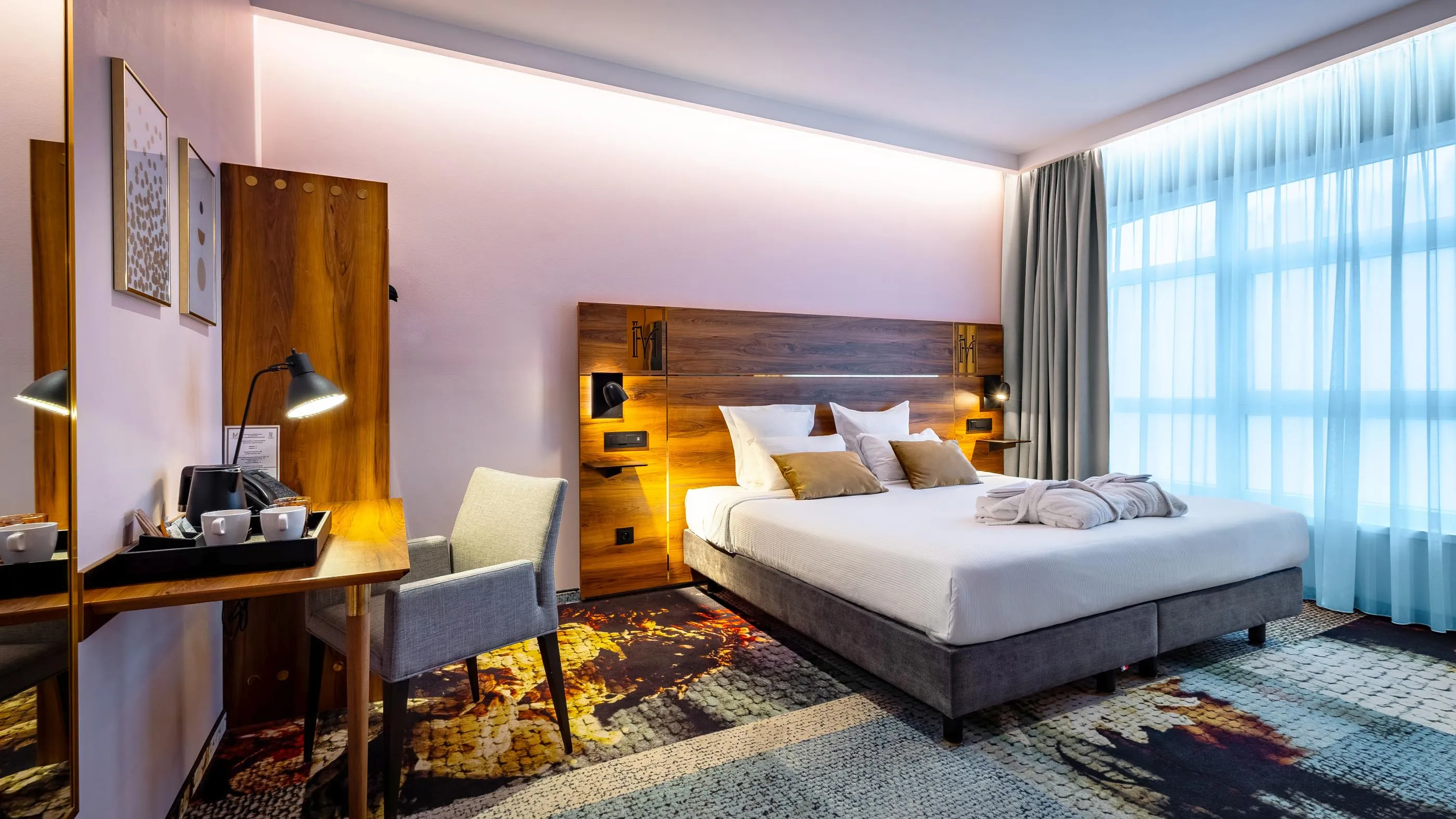 Chambre d'hôtel avec un lit double, une table de travail, une chaise et de grandes fenêtres laissant entrer la lumière du jour
