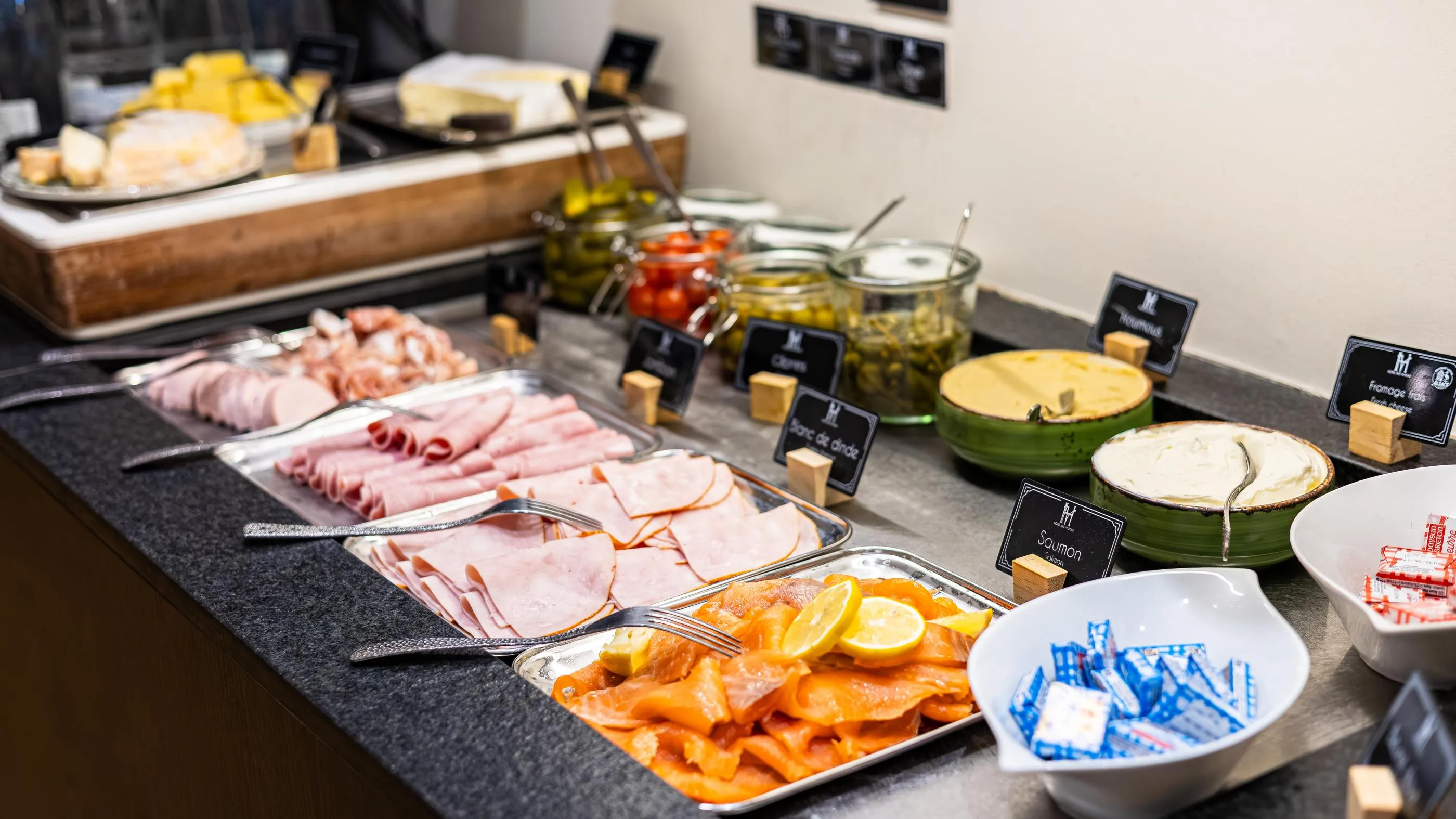 Un buffet avec divers plats de charcuterie, fromages, légumes marinés et saumon fumé, disposés sur une table avec des étiquettes en bois