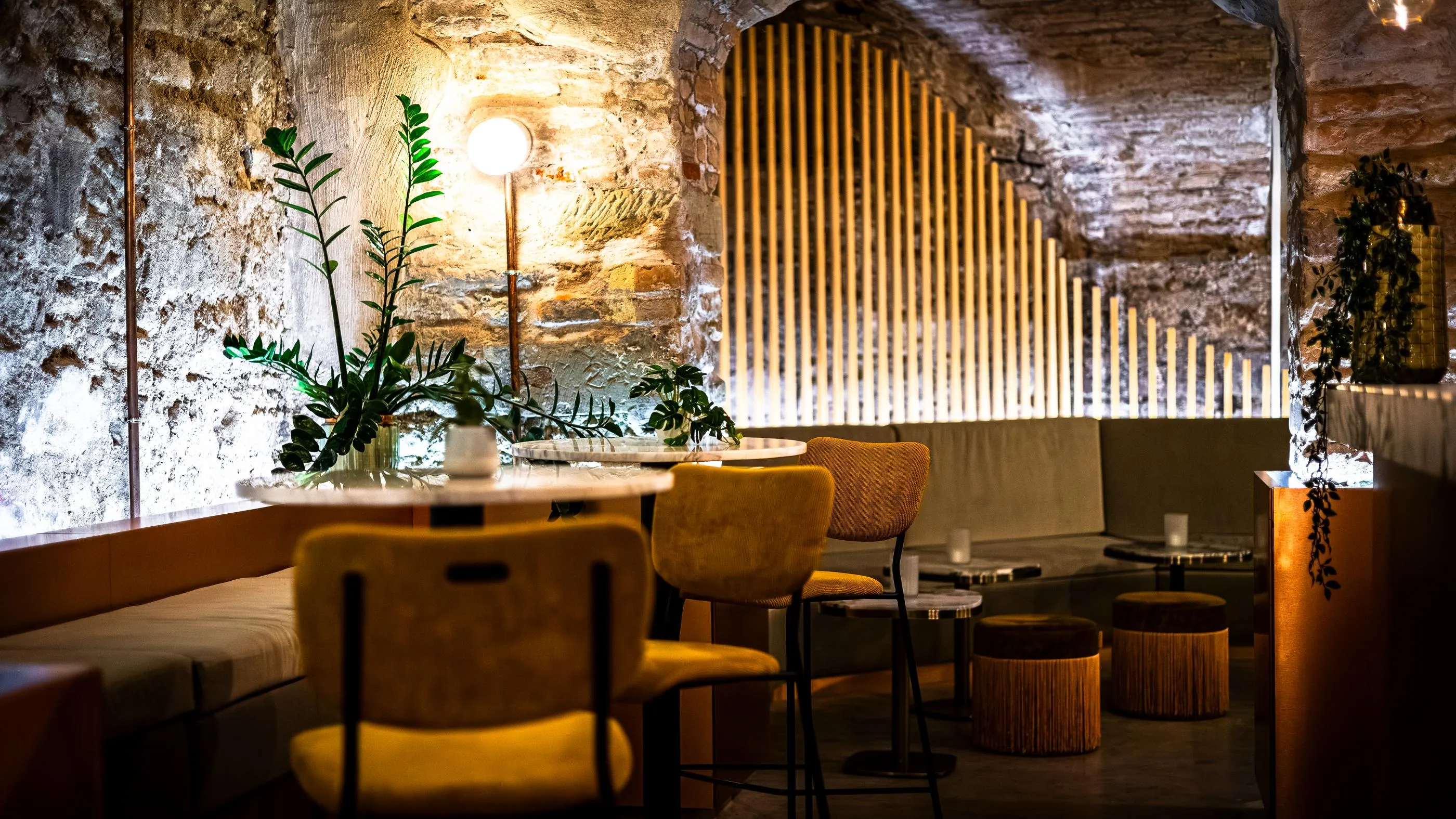 Un intérieur de café avec des murs en pierre, des tables, des chaises et des banquettes, éclairé par des bougies et des plantes vertes