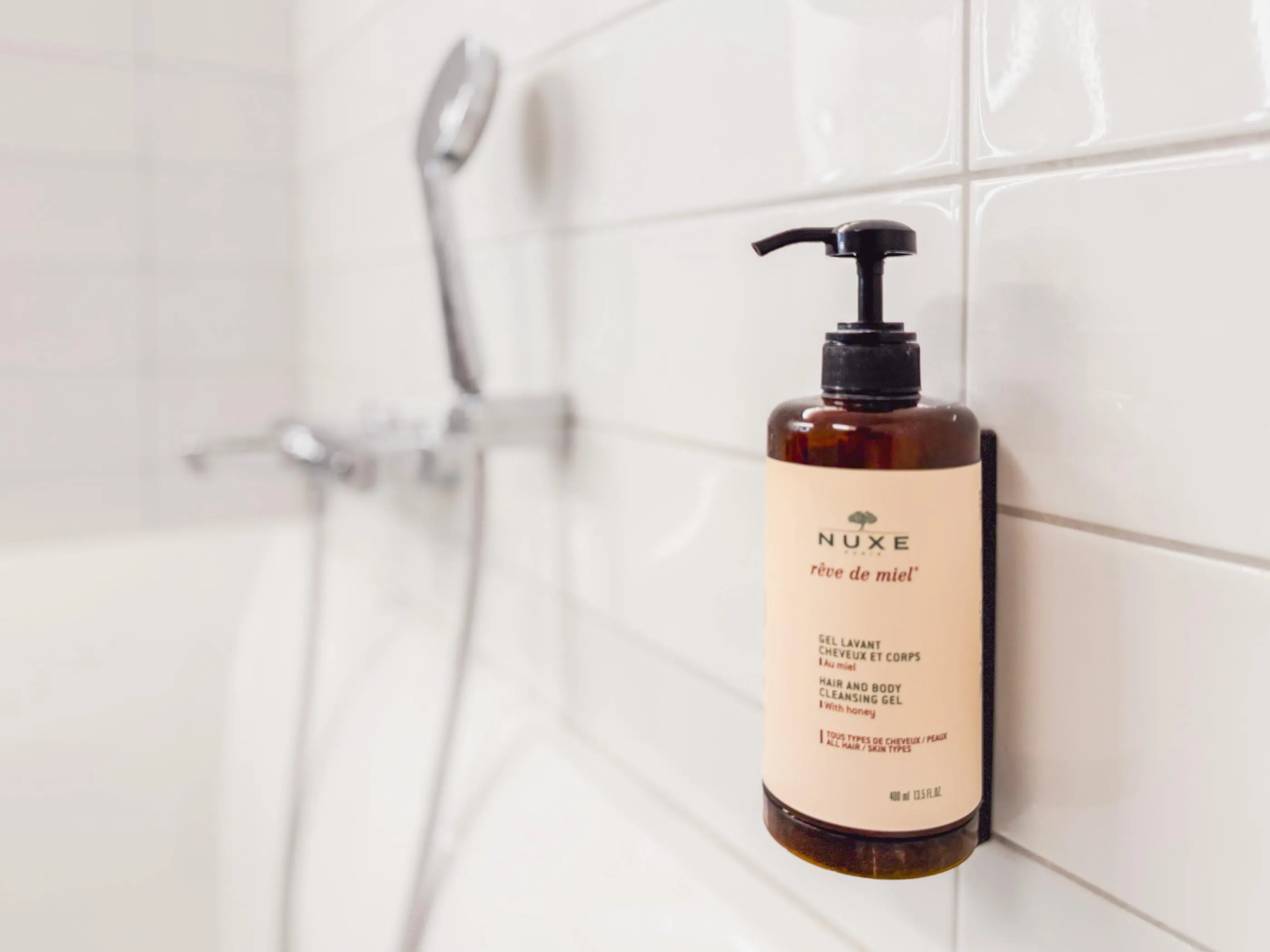Bouteille de shampoing Nuxe "rêve de miel" fixée sur un mur carrelé près d'un pommeau de douche