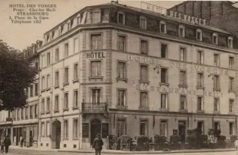 Ancien bâtiment de l'Hôtel des Vosges avec une personne marchant devant, des véhicules stationnés et des enseignes visibles sur la façade