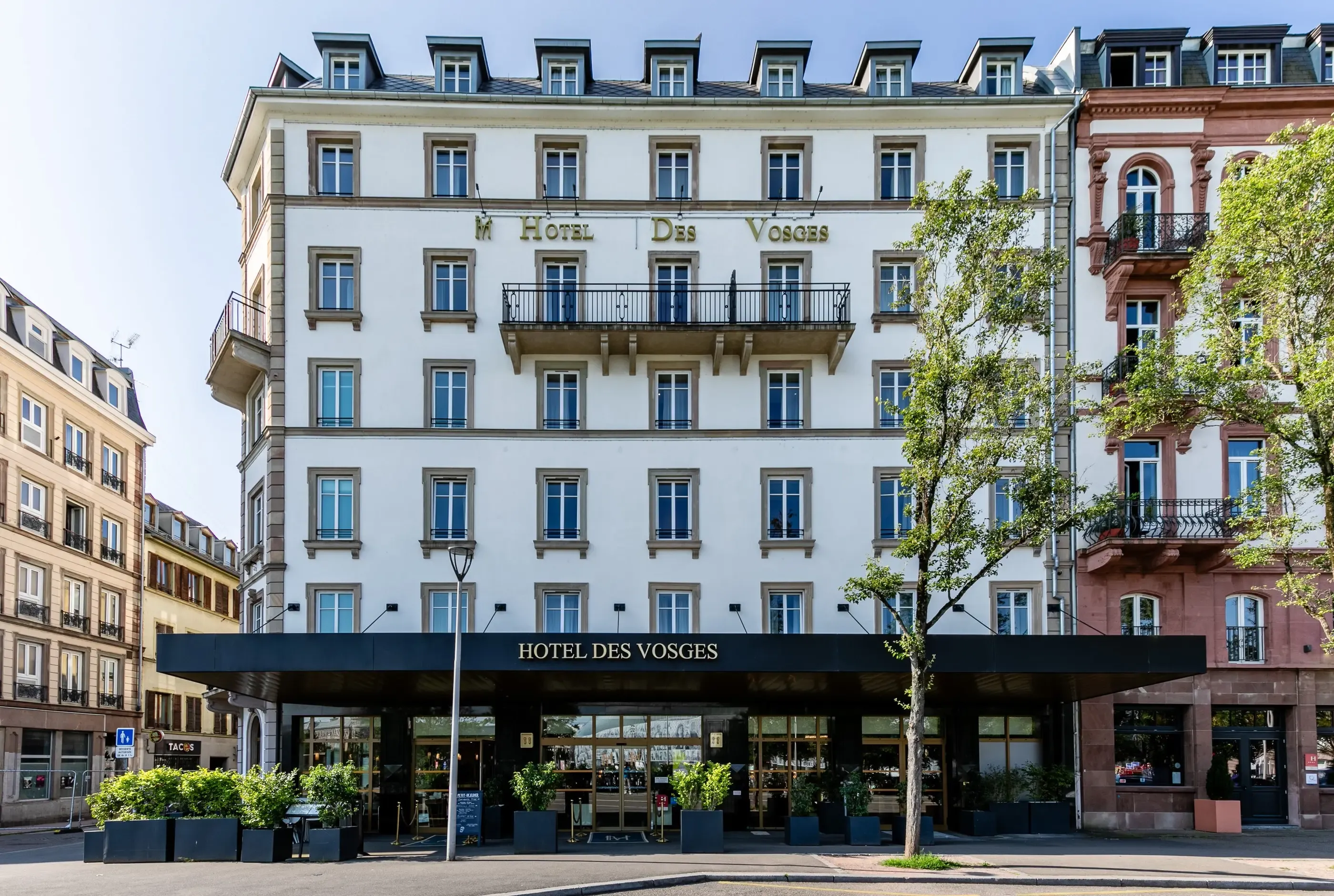 Hôtel des Vosges, bâtiment blanc à cinq étages avec balcons, situé à l'angle d'une rue avec des arbres et des plantes en pot devant l'entrée