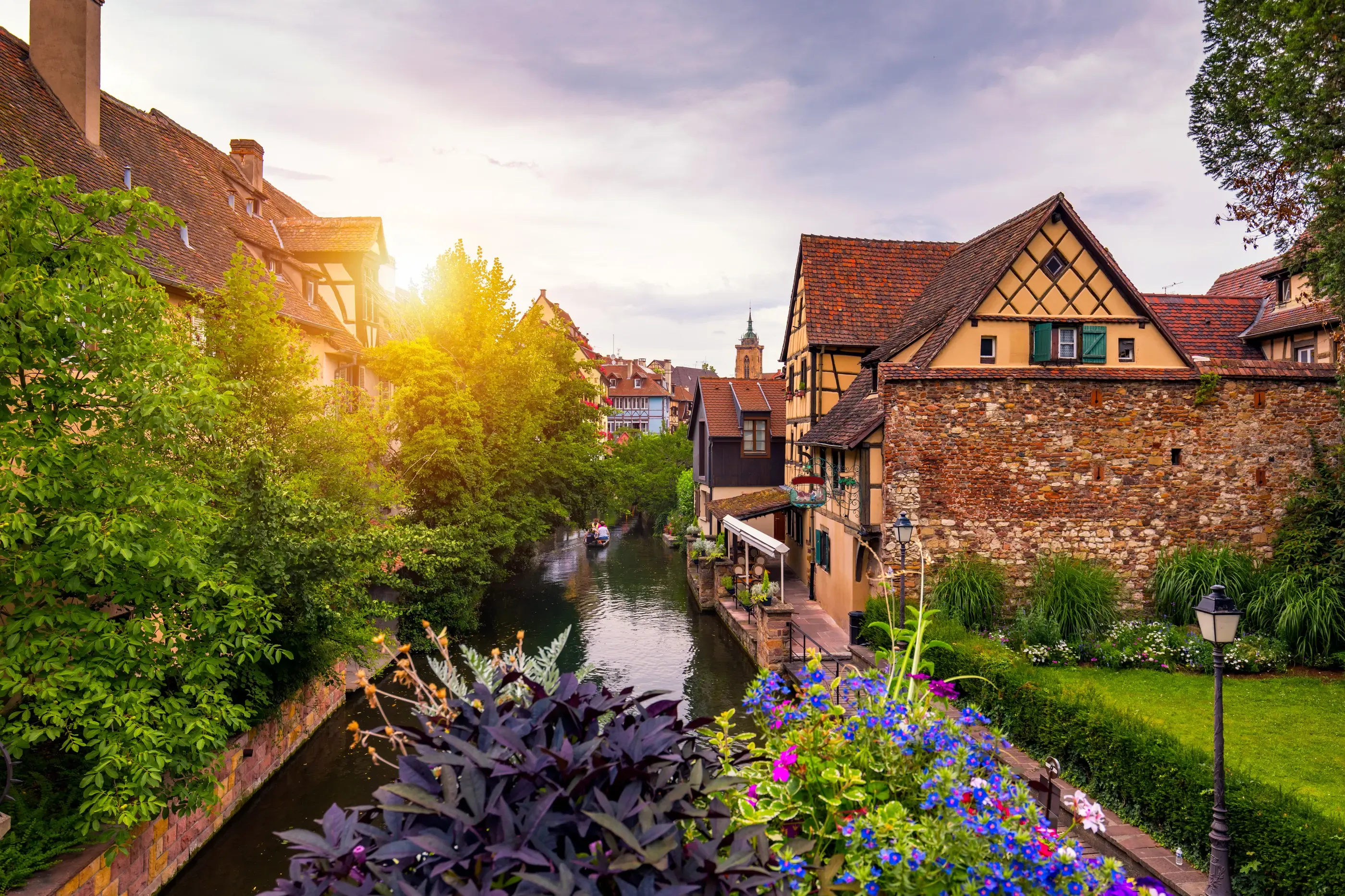 Un canal bordé de maisons à colombages et de végétation, avec des fleurs en premier plan et un ciel nuageux