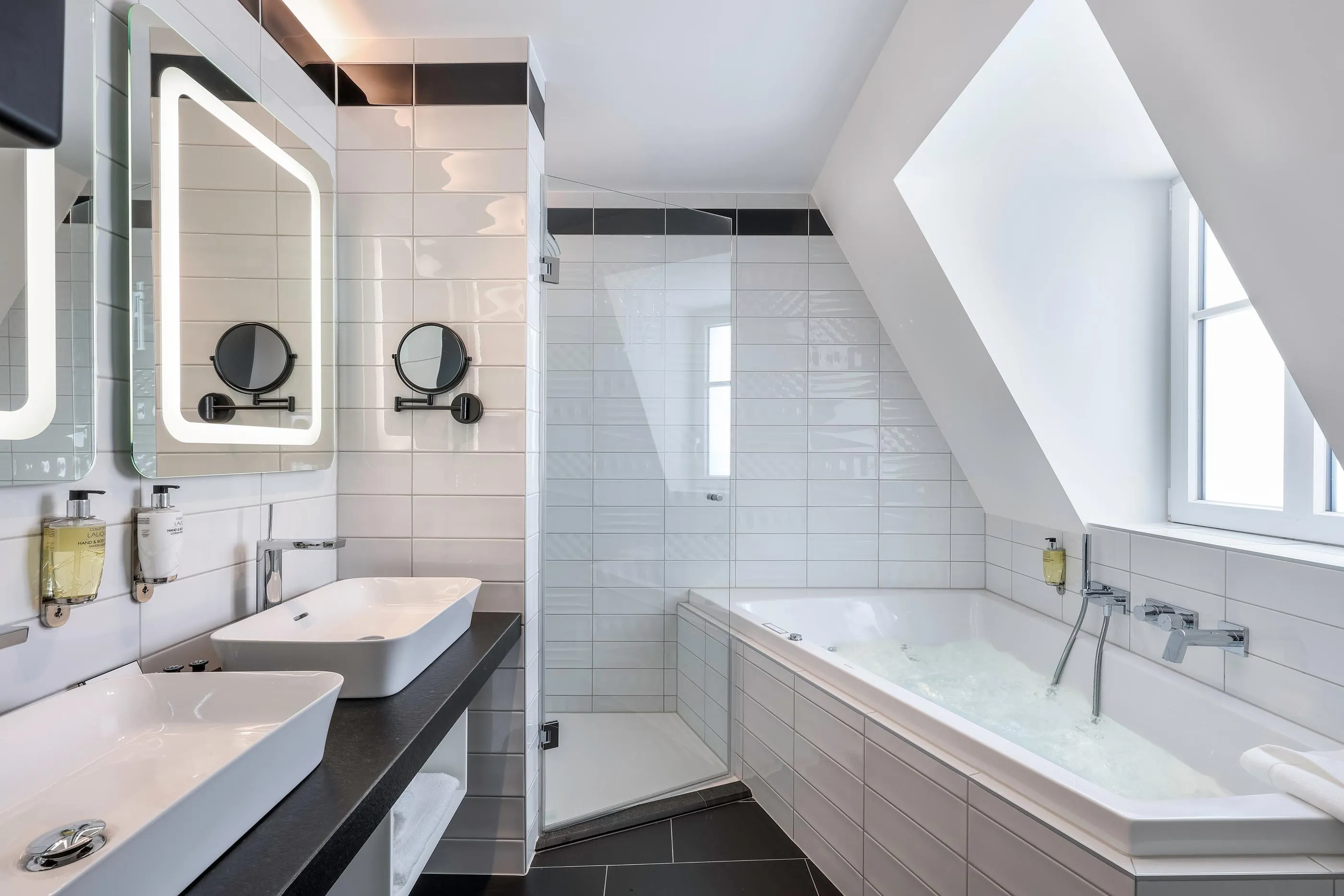Salle de bain moderne avec deux lavabos, une baignoire remplie d'eau et une douche, le tout carrelé de blanc et noir