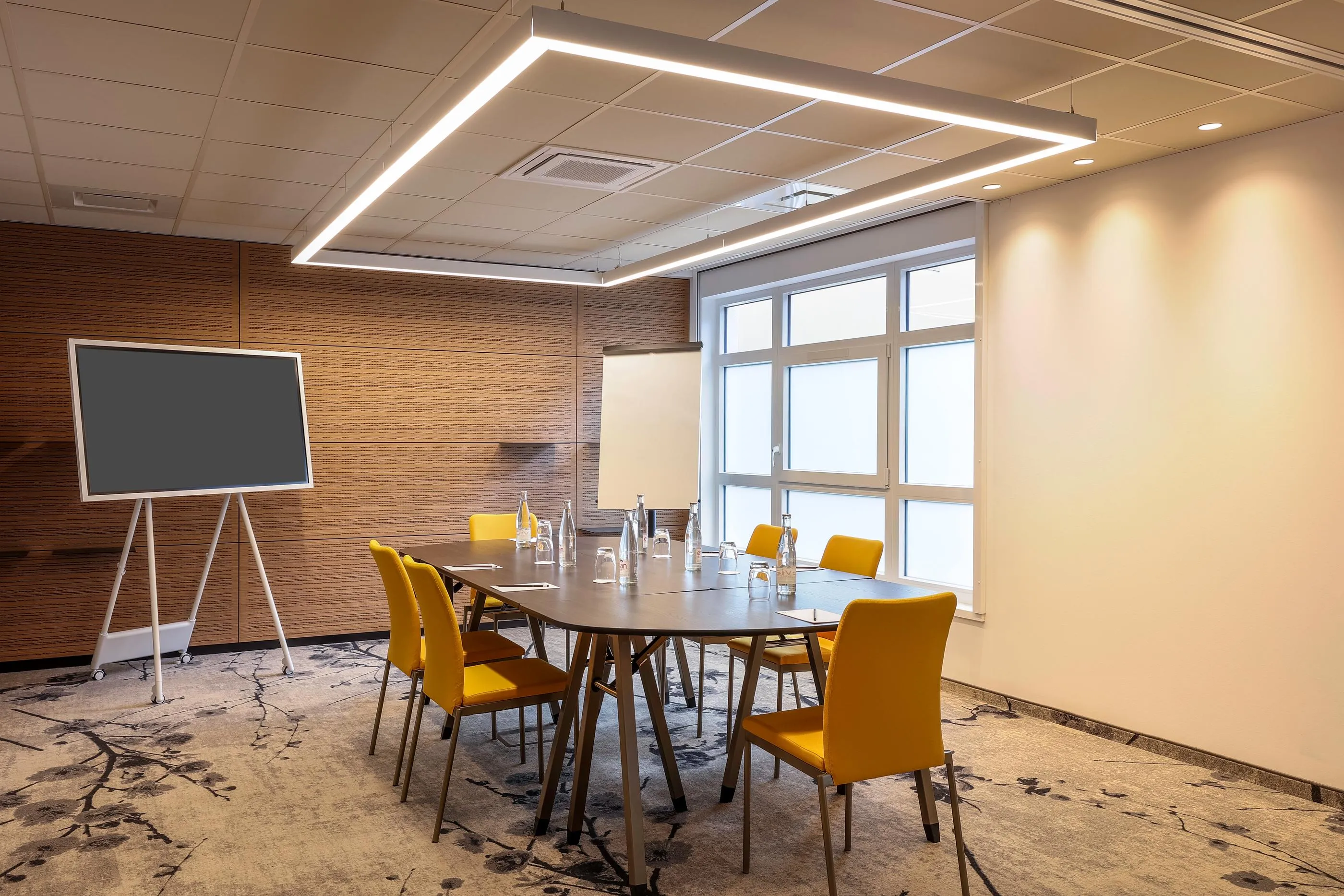 Salle de réunion avec table, chaises jaunes, écran et fenêtres