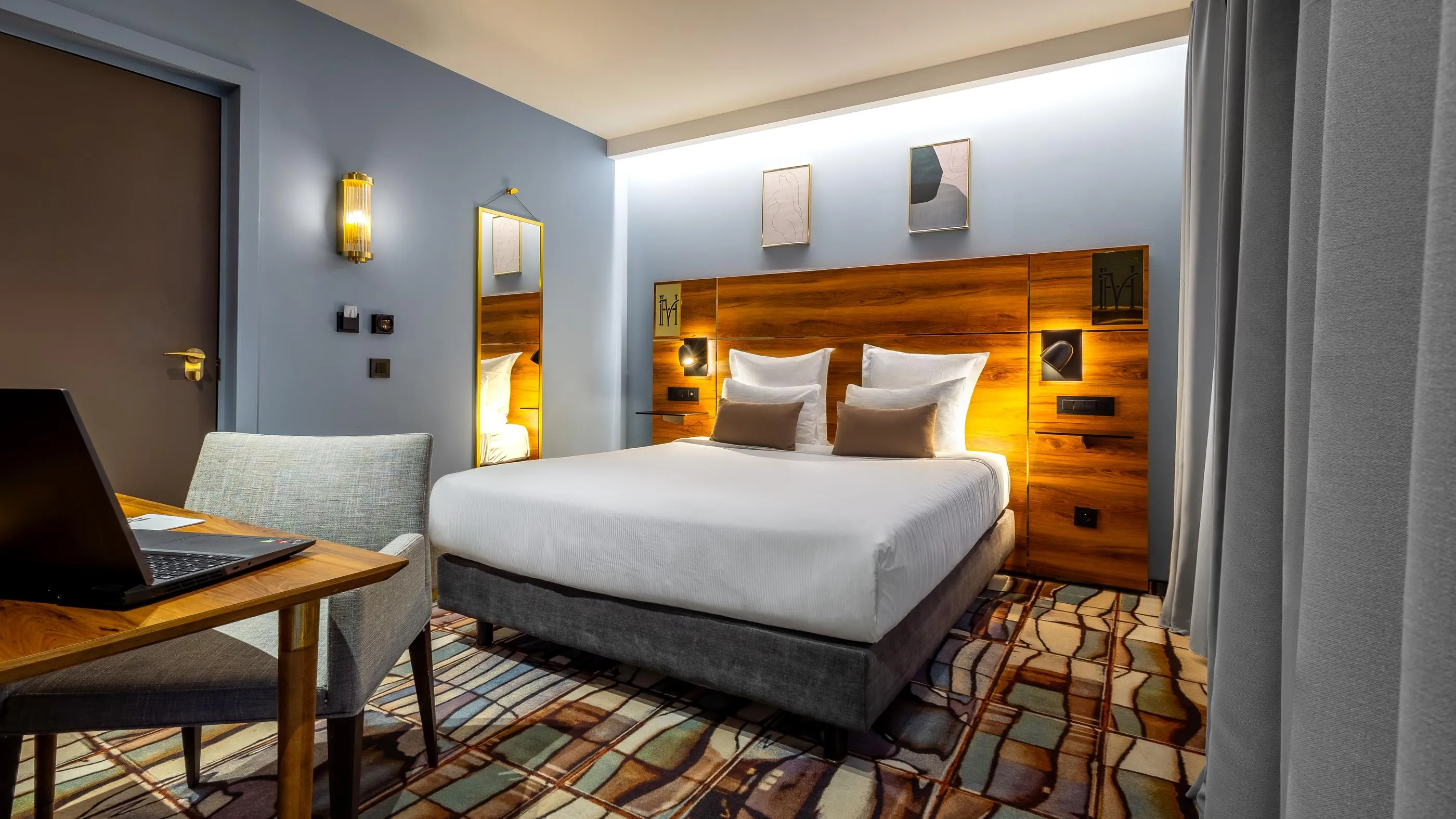 Chambre d'hôtel avec un lit double, une table de travail avec un ordinateur portable, un fauteuil et des œuvres d'art sur les murs