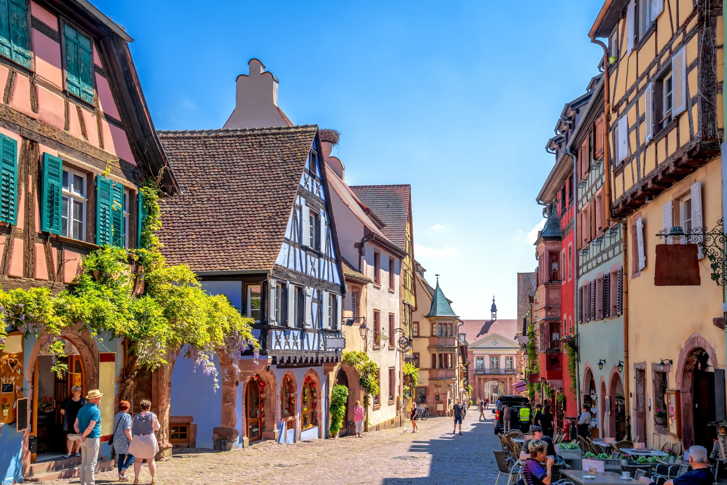 Rue pavée bordée de maisons à colombages colorées avec des gens qui marchent et des tables de café en terrasse