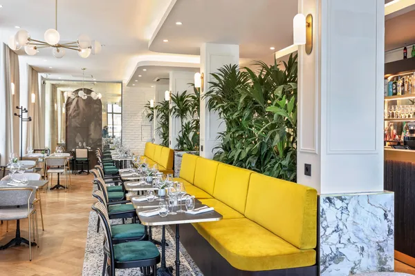 Restaurant élégant avec des tables dressées, des chaises vertes et une banquette jaune, des plantes vertes et un bar en marbre visible au fond