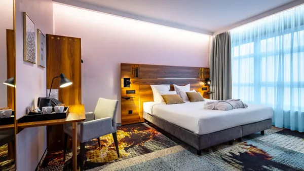Hotel room with a double bed, a work desk, a chair, and large windows letting in daylight.