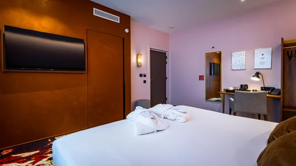 Hotel room with a bed covered in towels, a wall-mounted television, a desk with a lamp, and artwork on the wall.