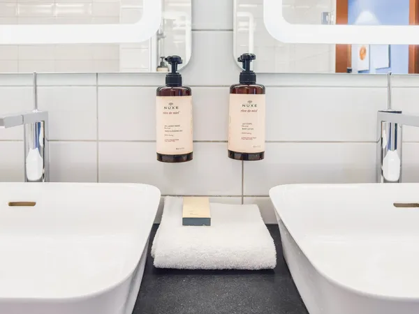 Deux distributeurs de produits Nuxe "rêve de miel" suspendus entre deux lavabos avec serviettes et savon